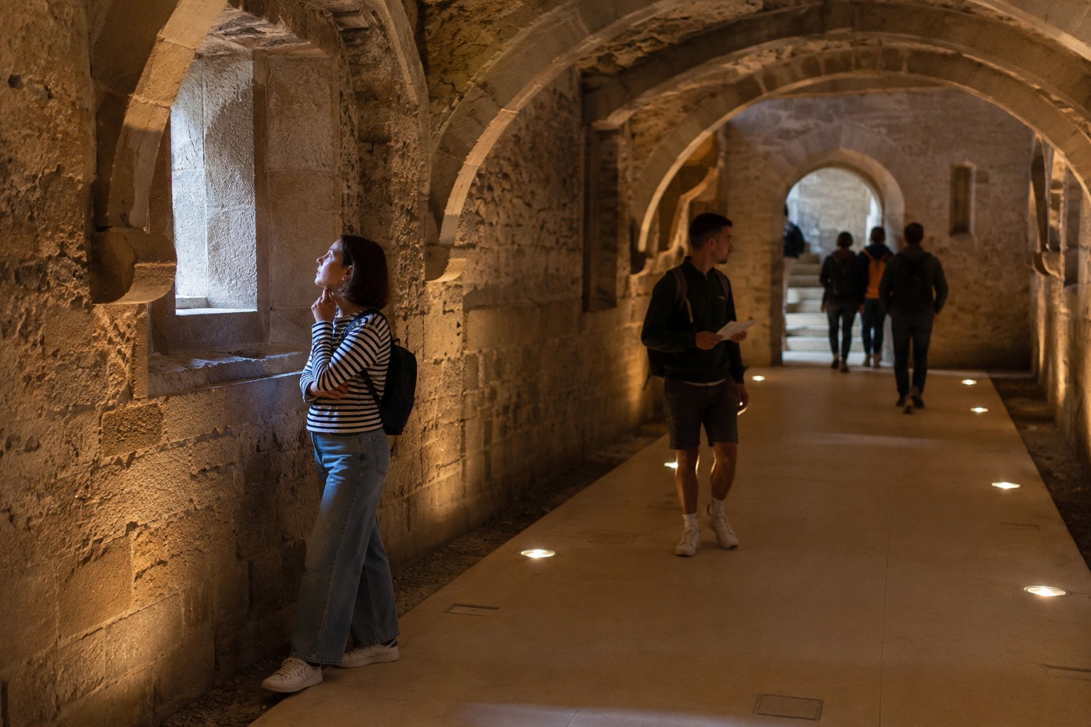 Visitante permanece observando en silencio un detalle del interior de un castillo mientras otros continúan el recorrido, reflejando distintas formas de vivir una misma experiencia cultural (imagen adaptada con IA)..